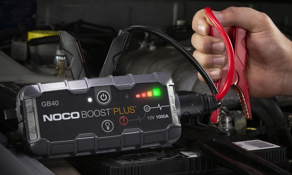 NOCO Boost Plus GB40 an einer Fahrzeugbatterie mit roter Batterieklemme in der Hand; keine Anschlussanleitung